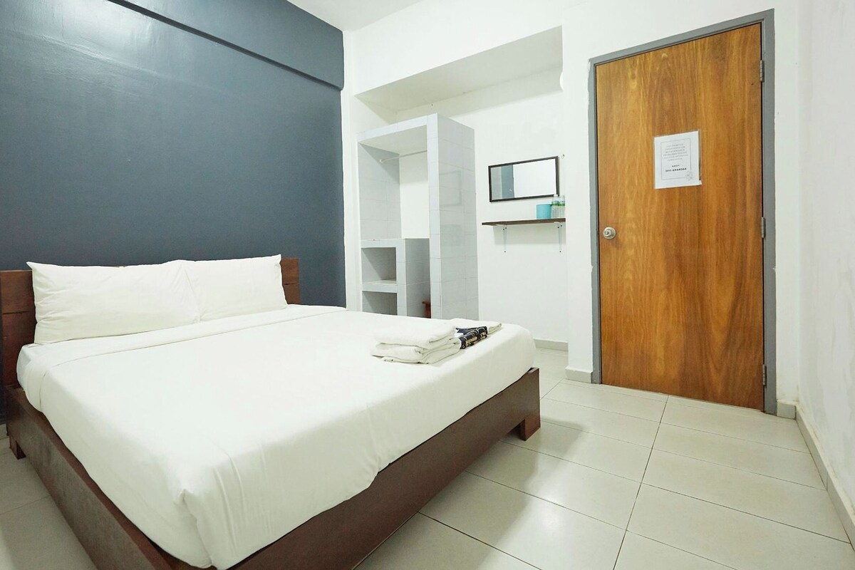 A neatly made bed with white linens, a wooden headboard, and a small bedside table with a lamp. A wooden door with a sign is visible in the background.
