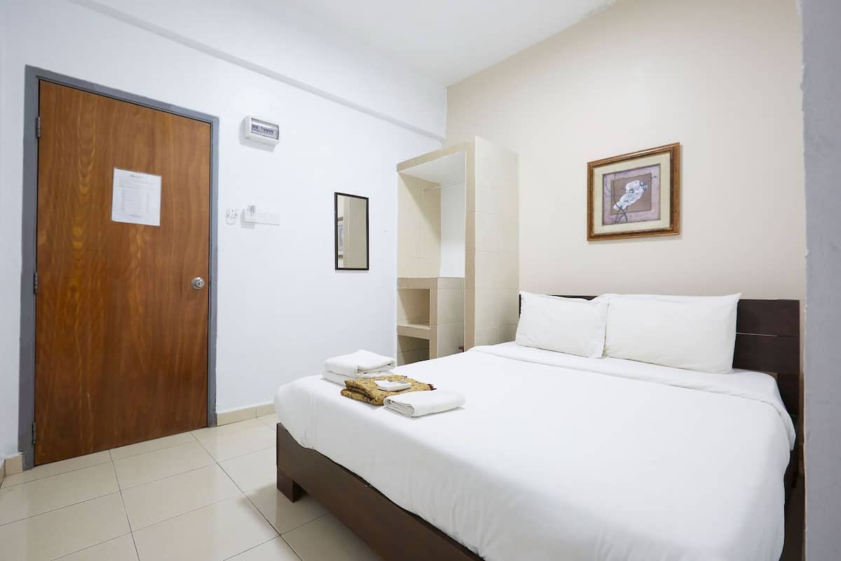 A hotel room with a wooden door, a bed with white linens, a framed picture on the wall, and a small shelf with toiletries.