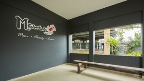 A room with a dark wall featuring a logo and the words "Peace, Family, Leisure." A wooden bench is placed against the wall, and a window reveals a view of greenery outside. - Mawar Villa (Kuantan)