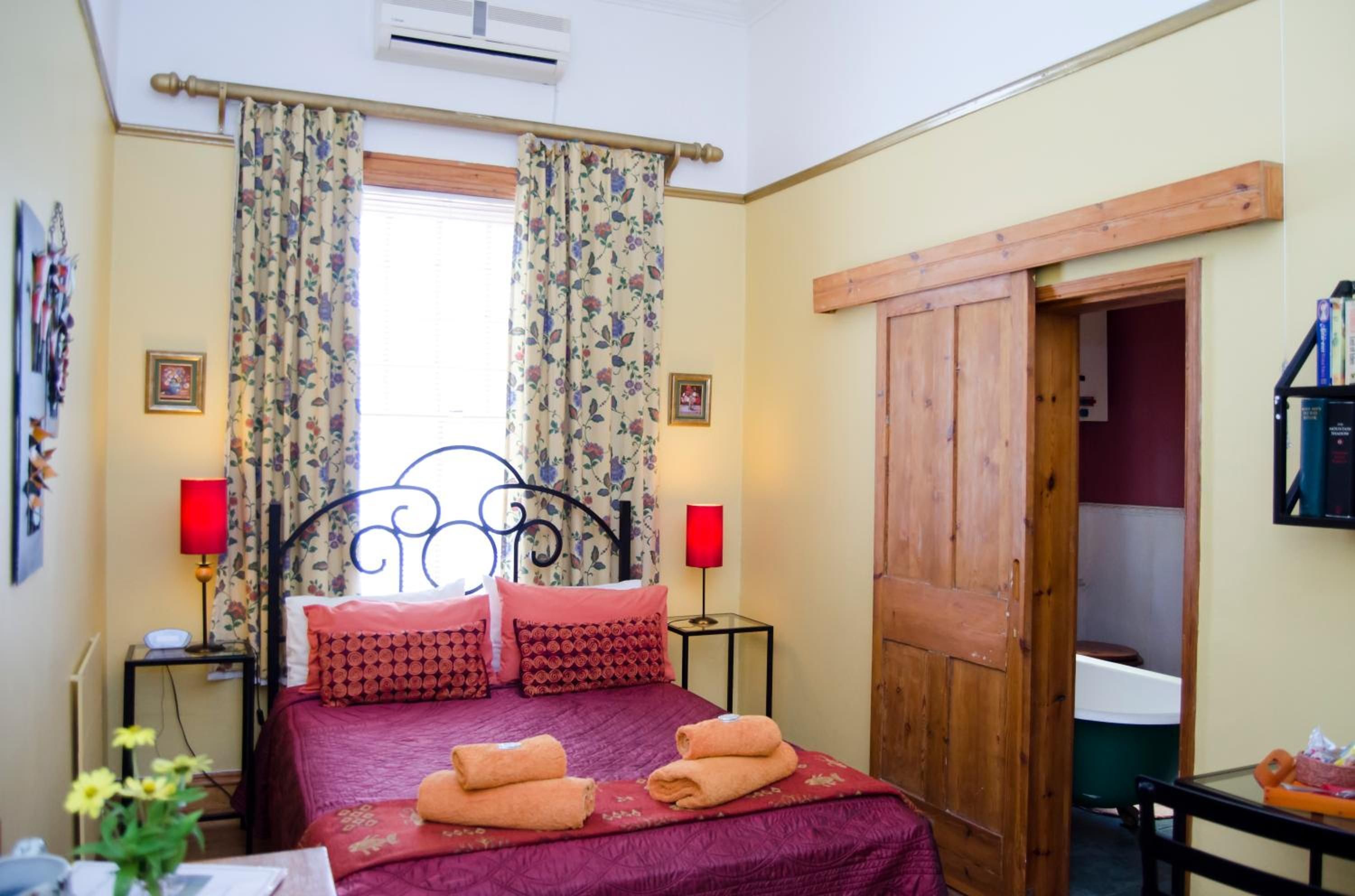 A bedroom with a bed, two bedside lamps, a window with floral curtains, and a wooden door.