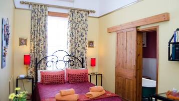 A bedroom with a bed, two bedside lamps, a window with floral curtains, and a wooden door.