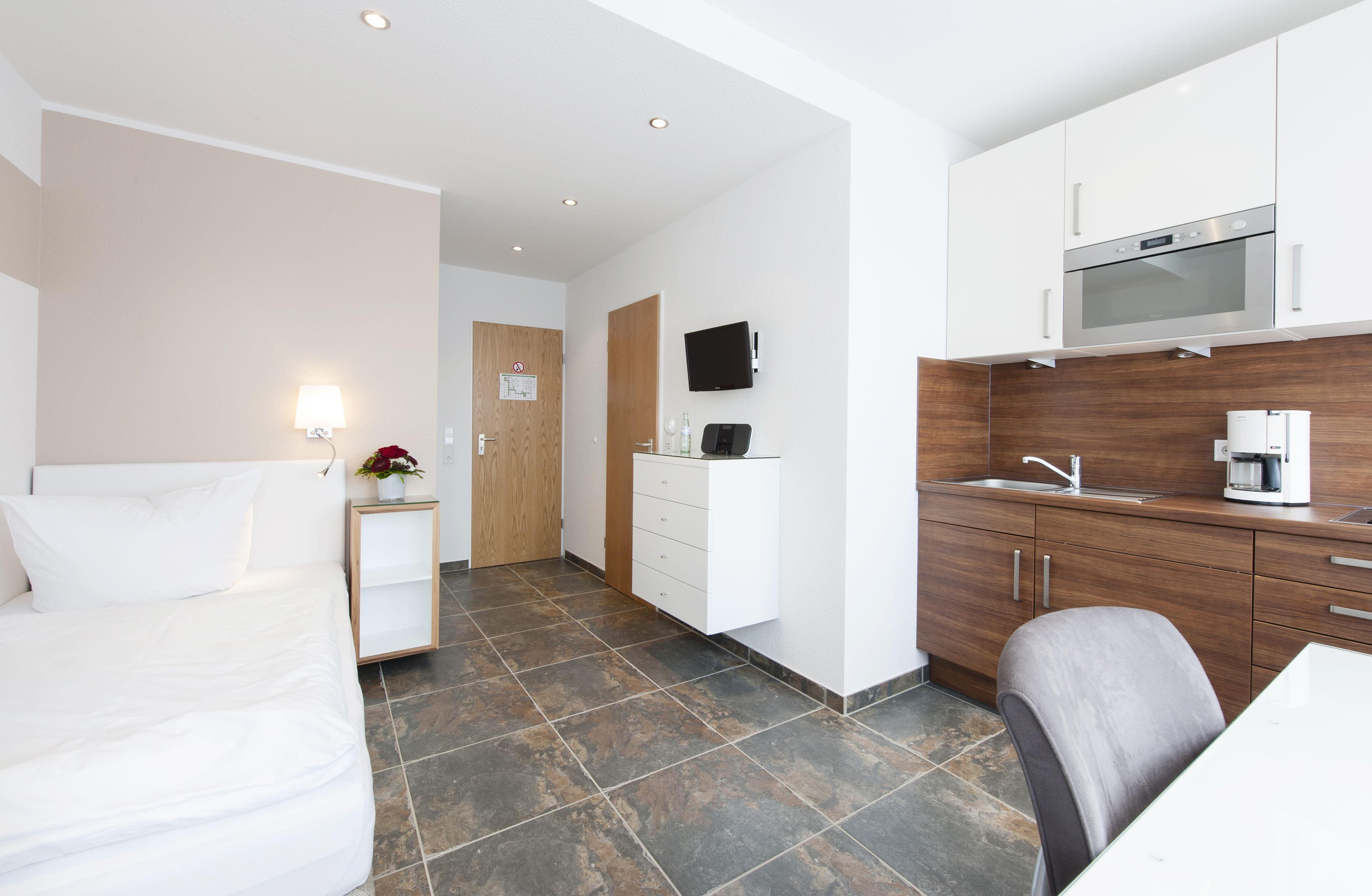 A modern hotel room with a bed, a small bedside table with a lamp, a wooden door, a kitchenette with a microwave, a coffee maker, and a TV on the wall.