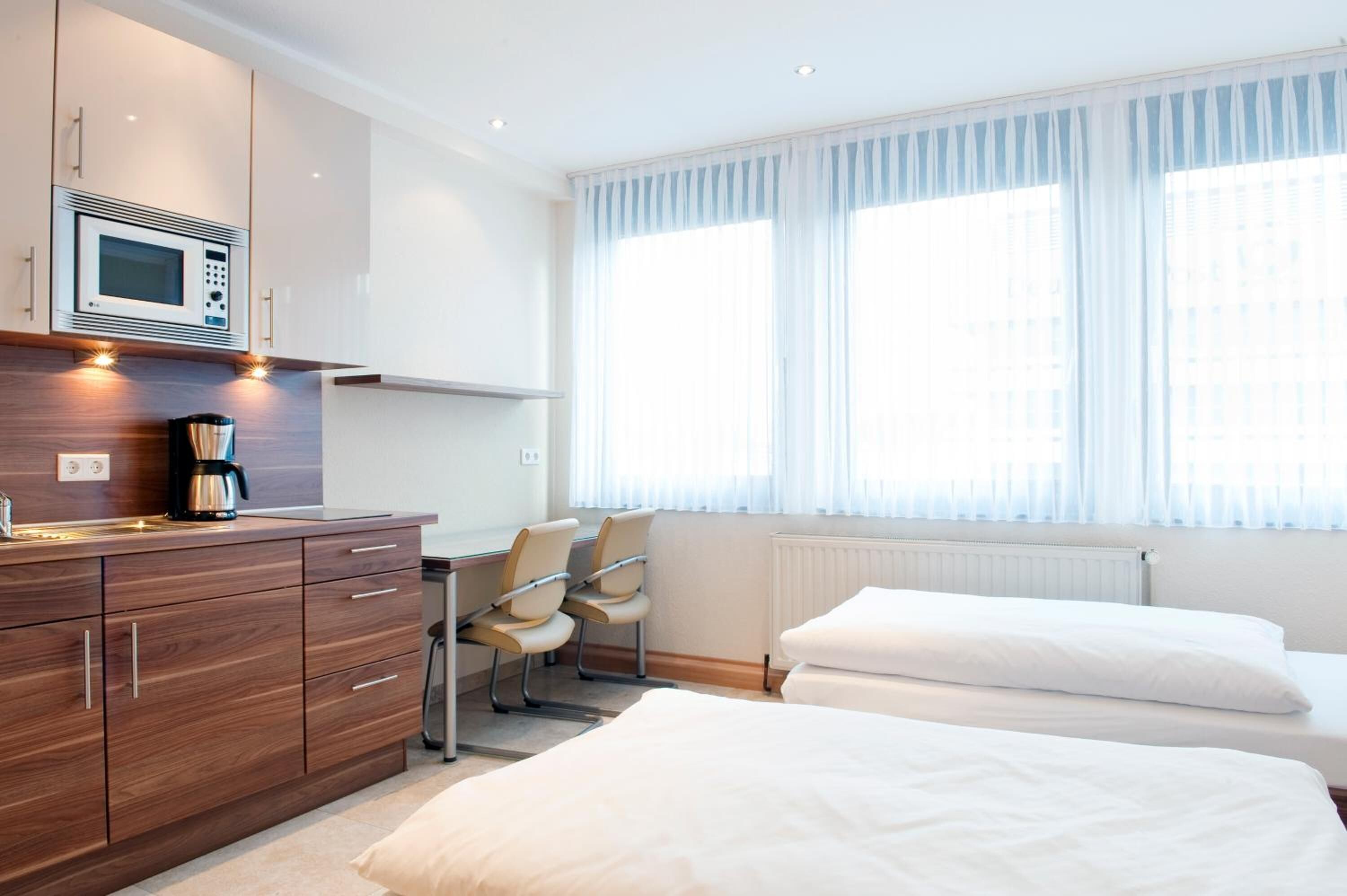 Une chambre d’hôtel équipée d’une kitchenette, d’un lit, d’un bureau avec des chaises et de grandes fenêtres avec des rideaux légers.