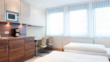 Une chambre d’hôtel équipée d’une kitchenette, d’un lit, d’un bureau avec des chaises et de grandes fenêtres avec des rideaux légers.