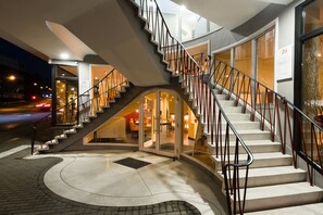 A modern hotel entrance with a curved staircase, glass doors, and a well-lit interior.