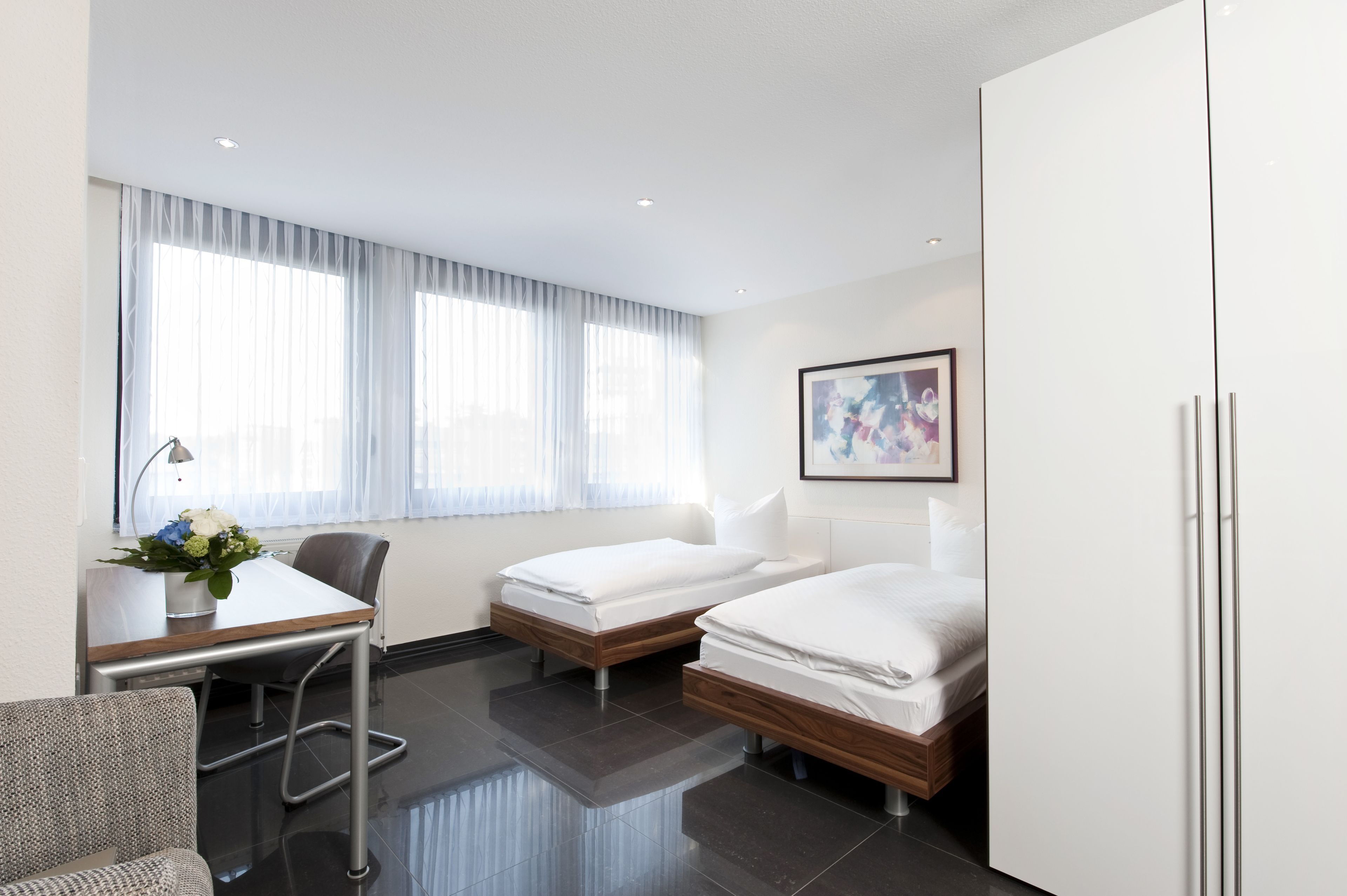 A hotel room with two single beds, a desk with a chair, a small table with a vase of flowers, and a large window with sheer curtains.