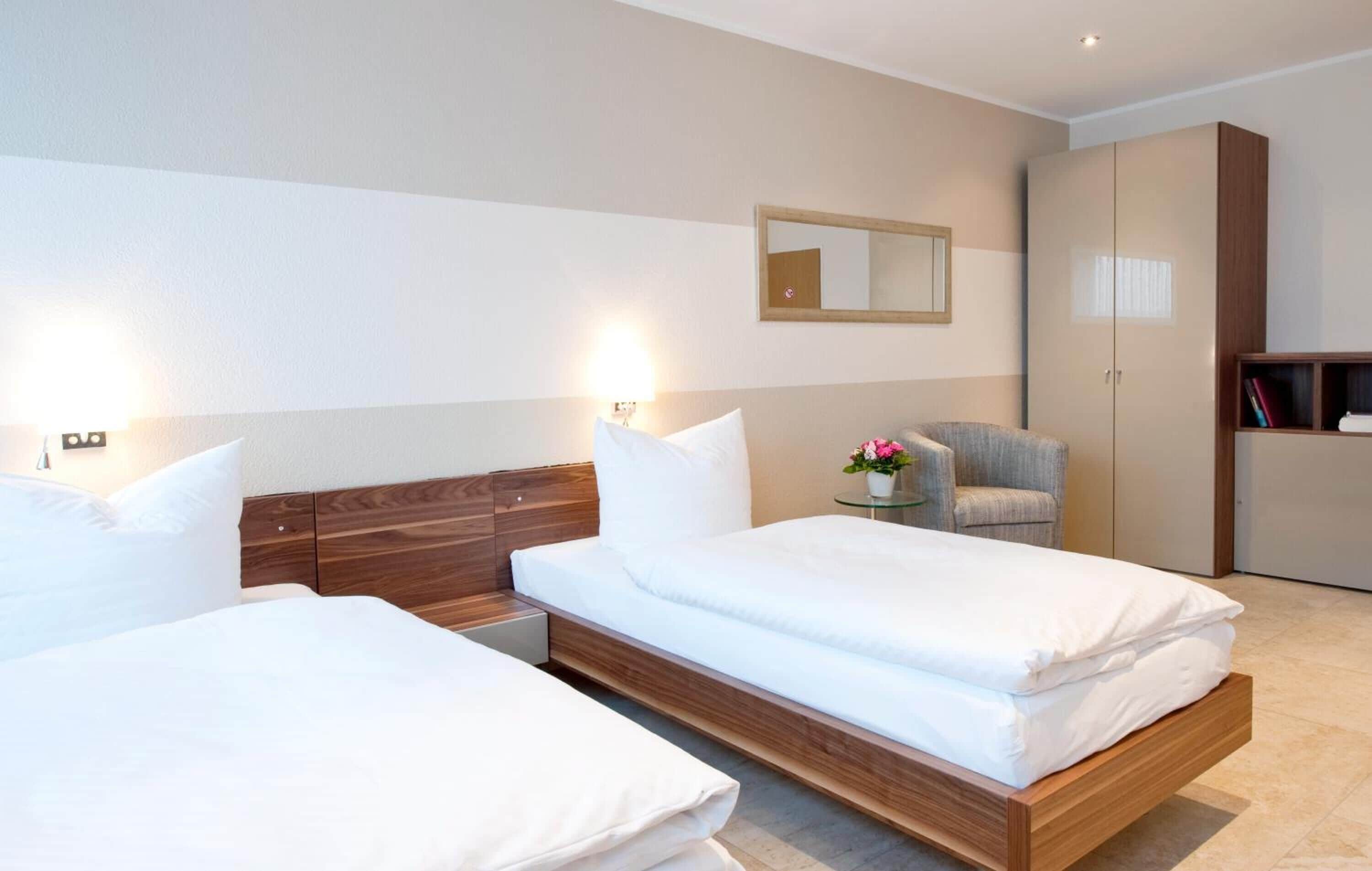 Ein modernes Hotelzimmer mit zwei Einzelbetten, einem kleinen runden Tisch mit einer Vase, einem Stuhl und einem Kleiderschrank.