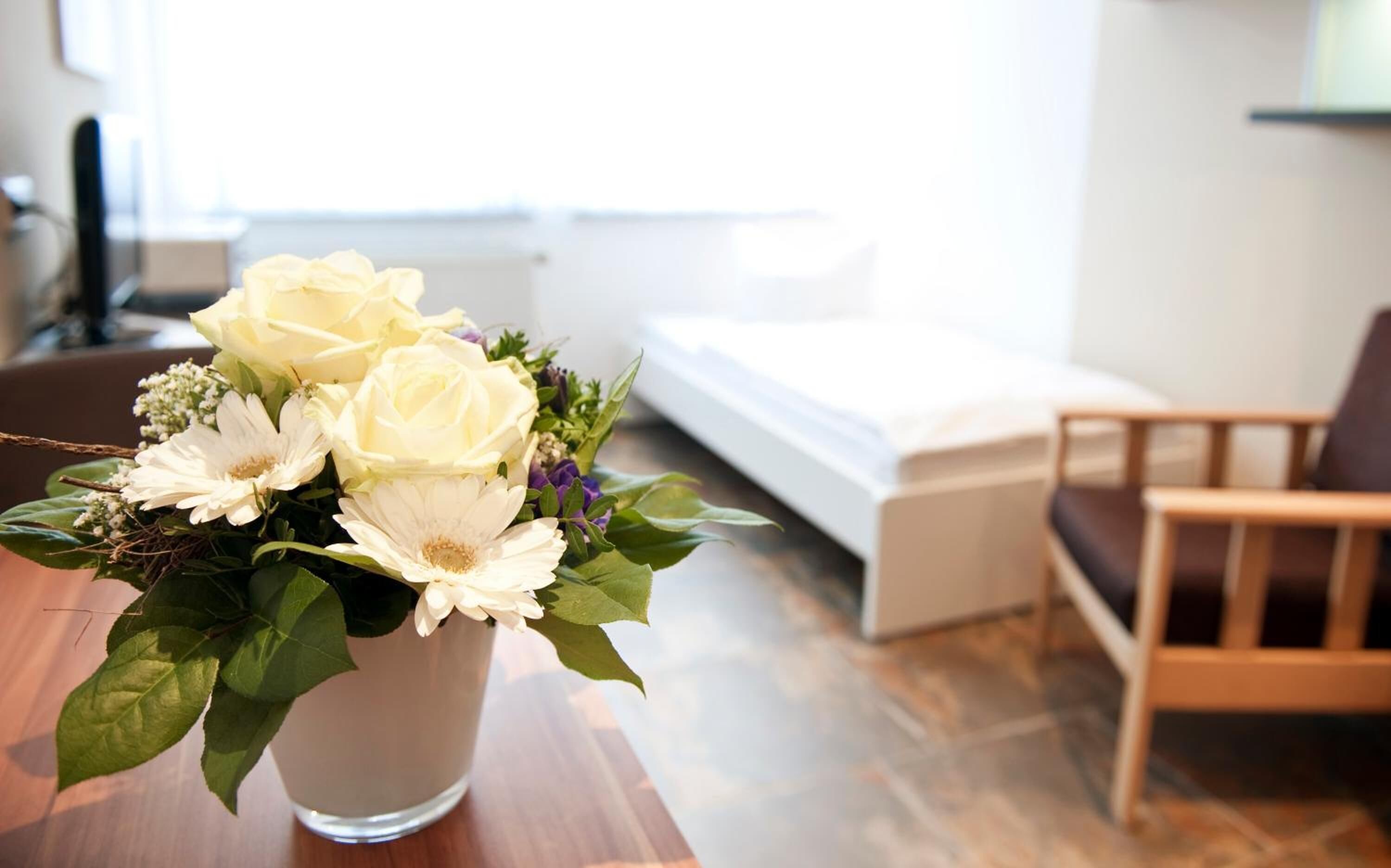 A room with a bed, a wooden chair, and a table with a vase of flowers.