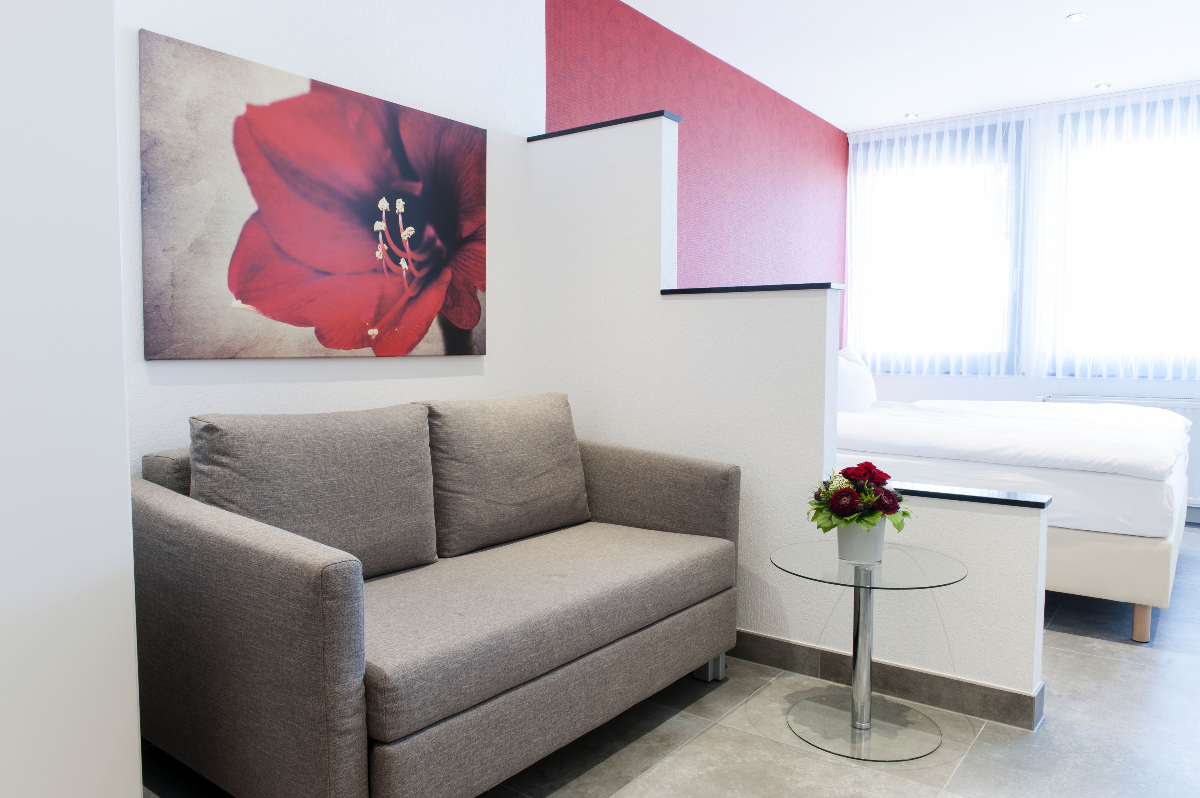 A modern room with a grey sofa, a small round table with a vase of flowers, and a large red flower painting on the wall.