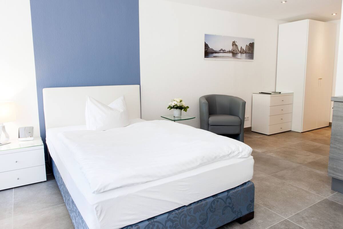 A modern bedroom with a large bed, a bedside table, a chair, a desk, and a wardrobe.