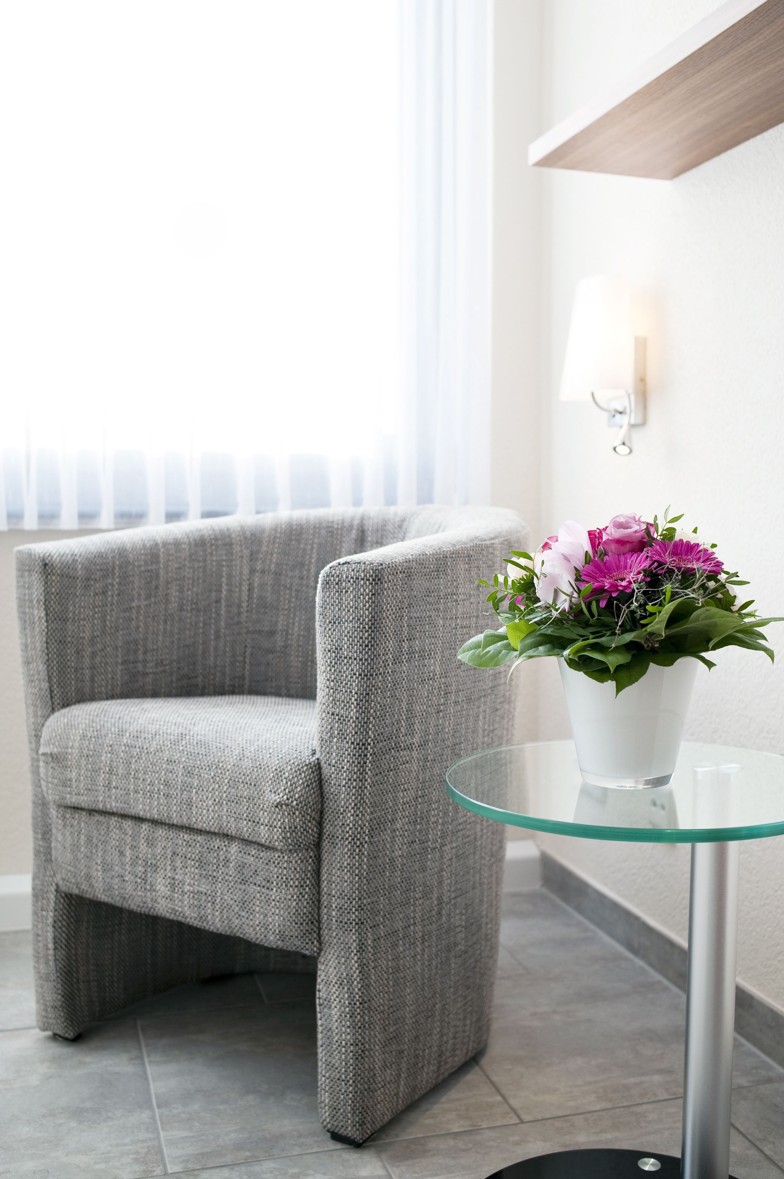 A grey armchair, a glass table with a flower vase, and a wall-mounted lamp.