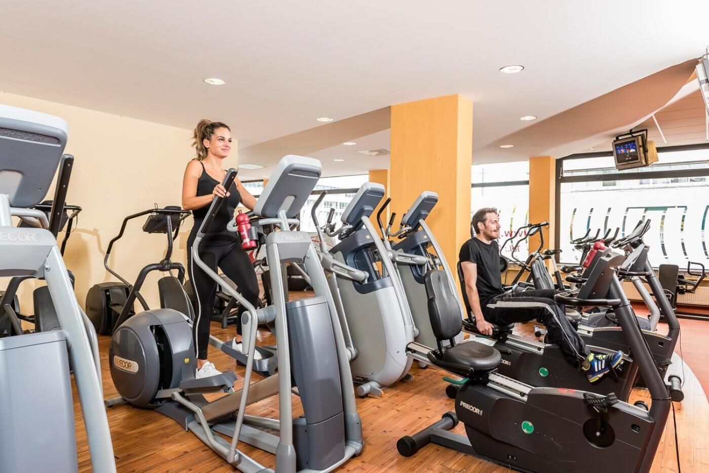 A gym with multiple treadmills and elliptical machines, wooden flooring, and a large window in the background.