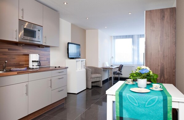 A modern kitchen with a microwave, coffee machine, and a flat-screen TV. There is a dining area with a table set for two, a chair, and a flower vase.