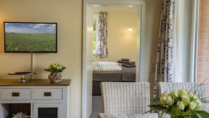 A room with a television on a stand, a small table with a plant, a wicker chair, and a bed with a view of a field through an open door. - Apartment-Hotel Dieksee Eck (Malente)