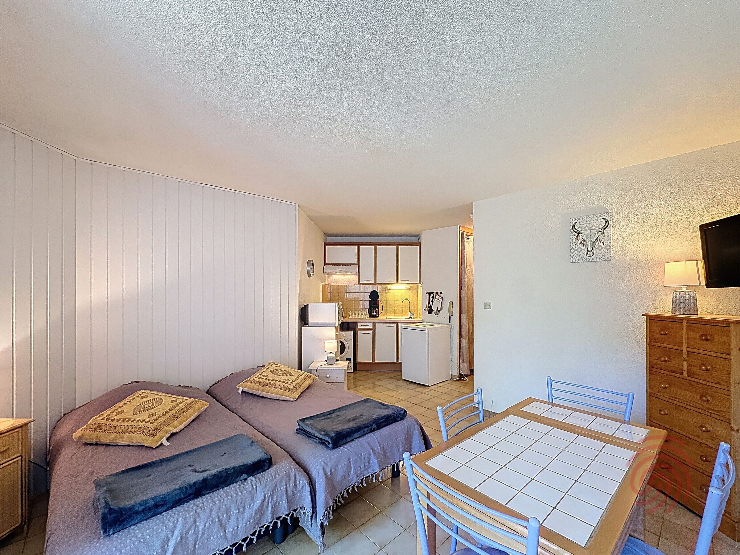 A compact room with two beds, a small kitchen, a dining table, and a TV.