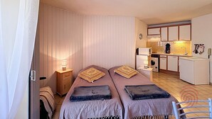 A bedroom with two beds, a nightstand, a lamp, and a kitchenette in the background. - Cozy Apartment with Terrace, Near Spa and Town Center, Pet-Friendly, Free Parking, 24m² (Lamalou-les-Bains)