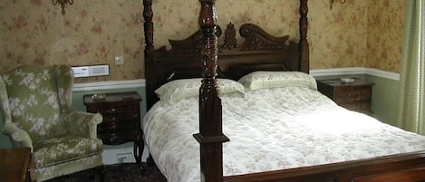Una cama con dosel con dosel adornada con un estampado floral, una mesita de noche de madera, un sillón tapizado color verde y una alfombra con motivos florales.