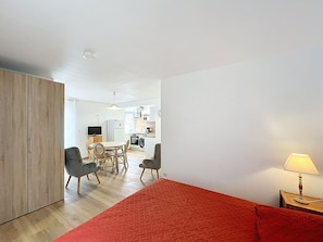 Una camera da letto con una coperta rossa, un armadio in legno, un angolo pranzo con sedie, una cucina con elettrodomestici e una lampada su un comodino.