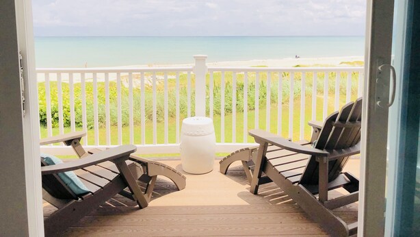 A balcony with two lounge chairs and a small table, overlooking a beach with a white fence and grassy area. - THE BEACH SHACK, a stylish oceanfront townhouse on quiet corner of Indialantic! (Indialantic)