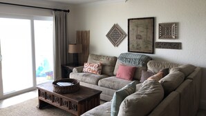 A living room with a sofa, a coffee table, and a lamp. There is a window with curtains and a view outside. - THE BEACH SHACK, a stylish oceanfront townhouse on quiet corner of Indialantic! (Indialantic)