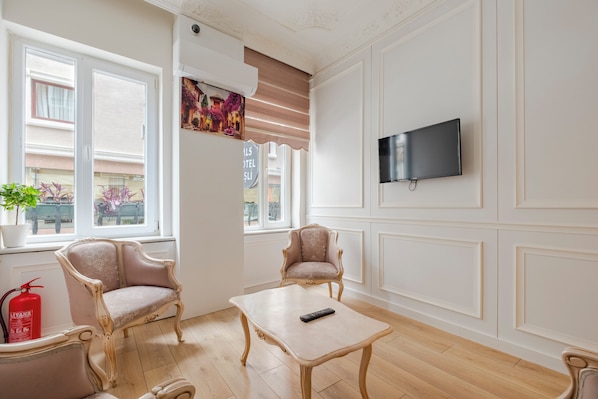 Une pièce comprenant deux fauteuils, une petite table, un téléviseur fixé au mur et une fenêtre avec des stores.
