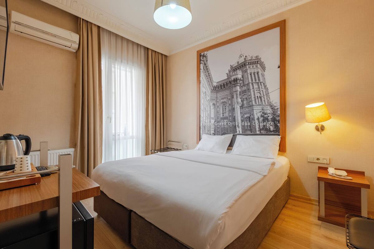 A hotel room with a large bed, a desk with a kettle, a lamp, and a framed picture on the wall.