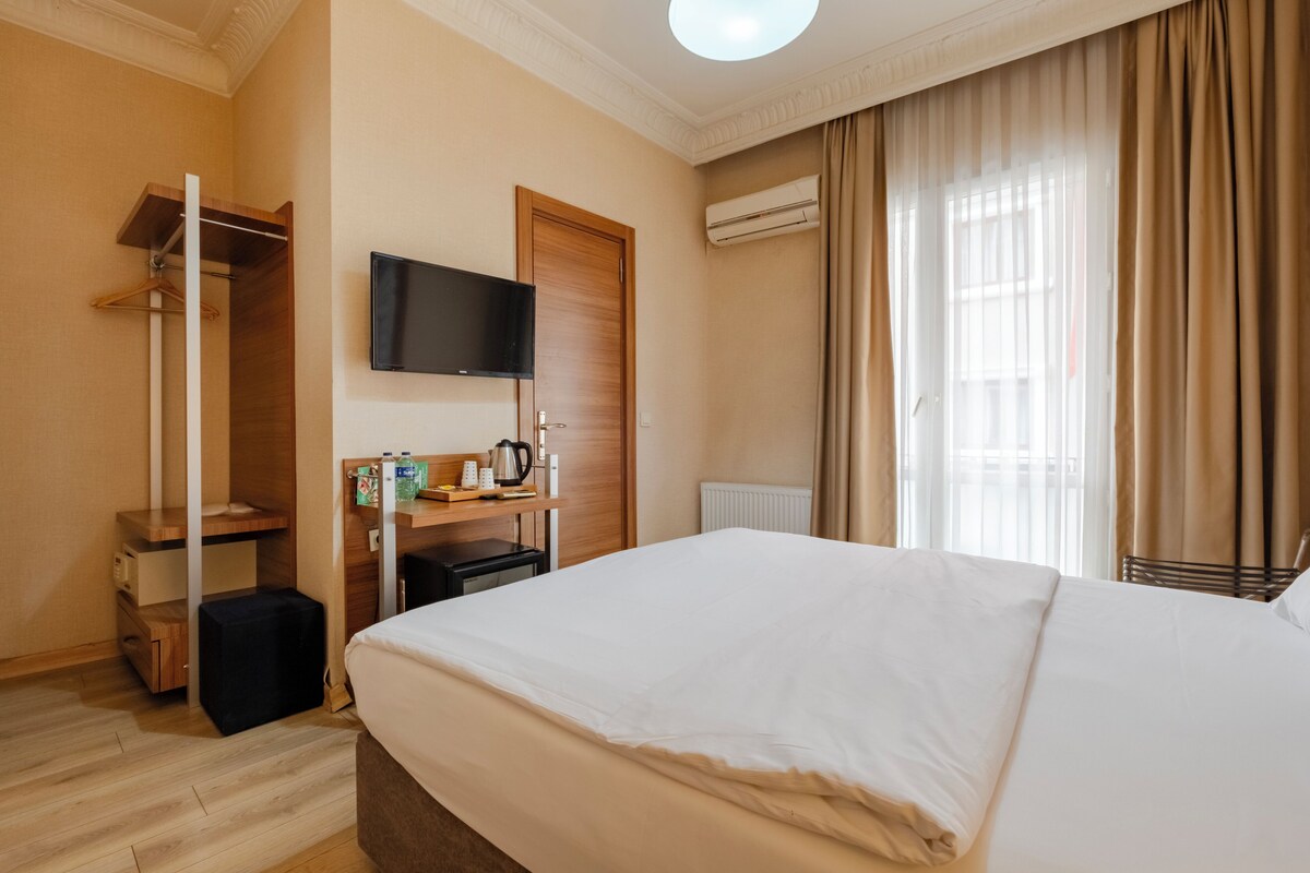 A hotel room with a large bed, a TV mounted on the wall, a wooden wardrobe, a desk with a kettle, and a radiator.