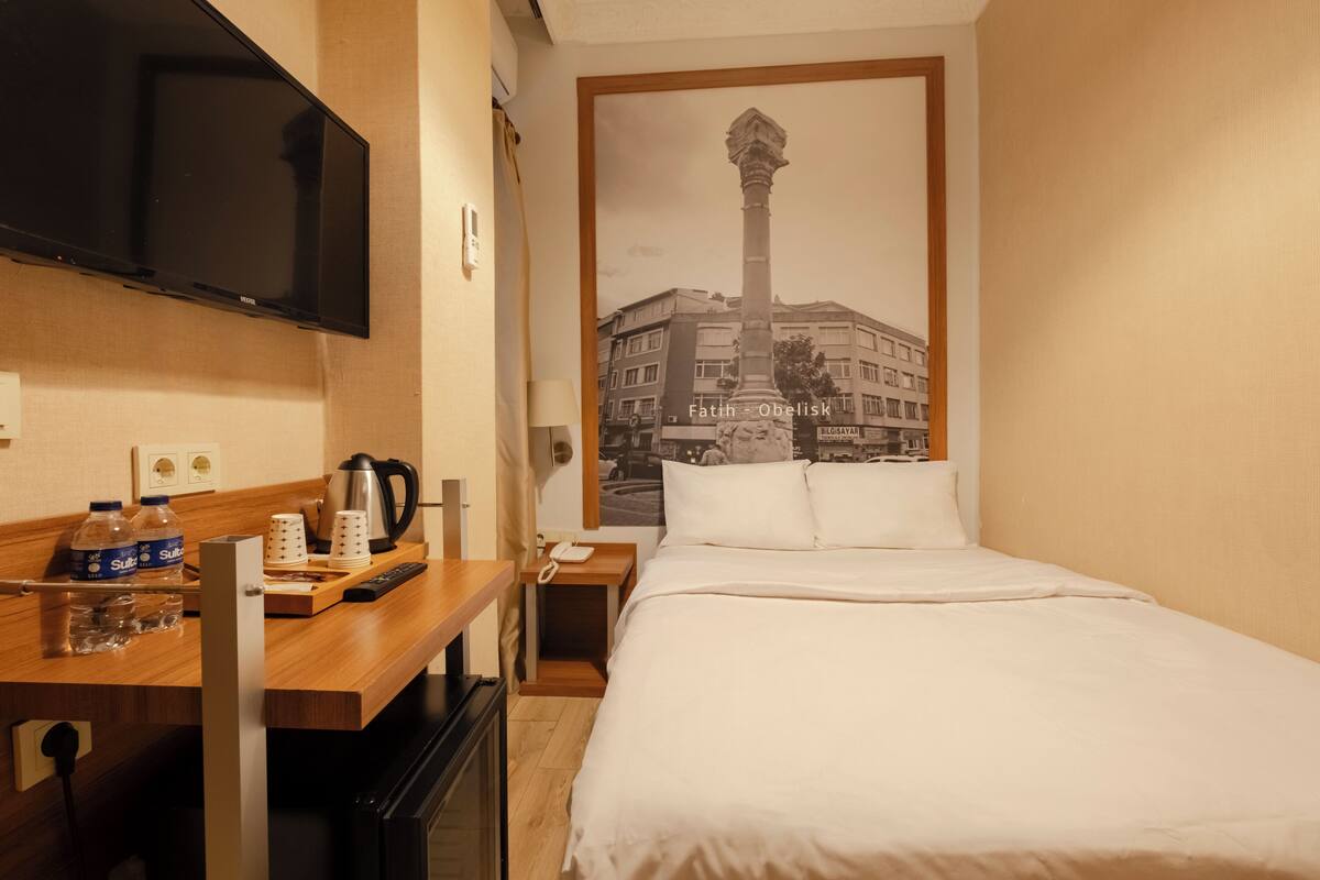 A hotel room with a bed, a TV mounted on the wall, a coffee maker, a telephone, and a poster of a building.