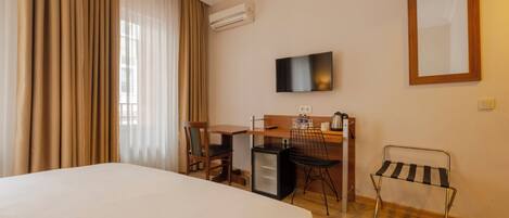 Quarto de hotel com cama, escrivaninha, cadeira, televisão e pequena geladeira.