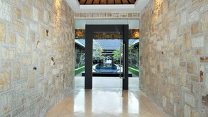 A hallway with stone walls and a tiled floor, leading to a glass door with a view of a garden and a building outside. - Villa Samudra Raya (Seminyak)