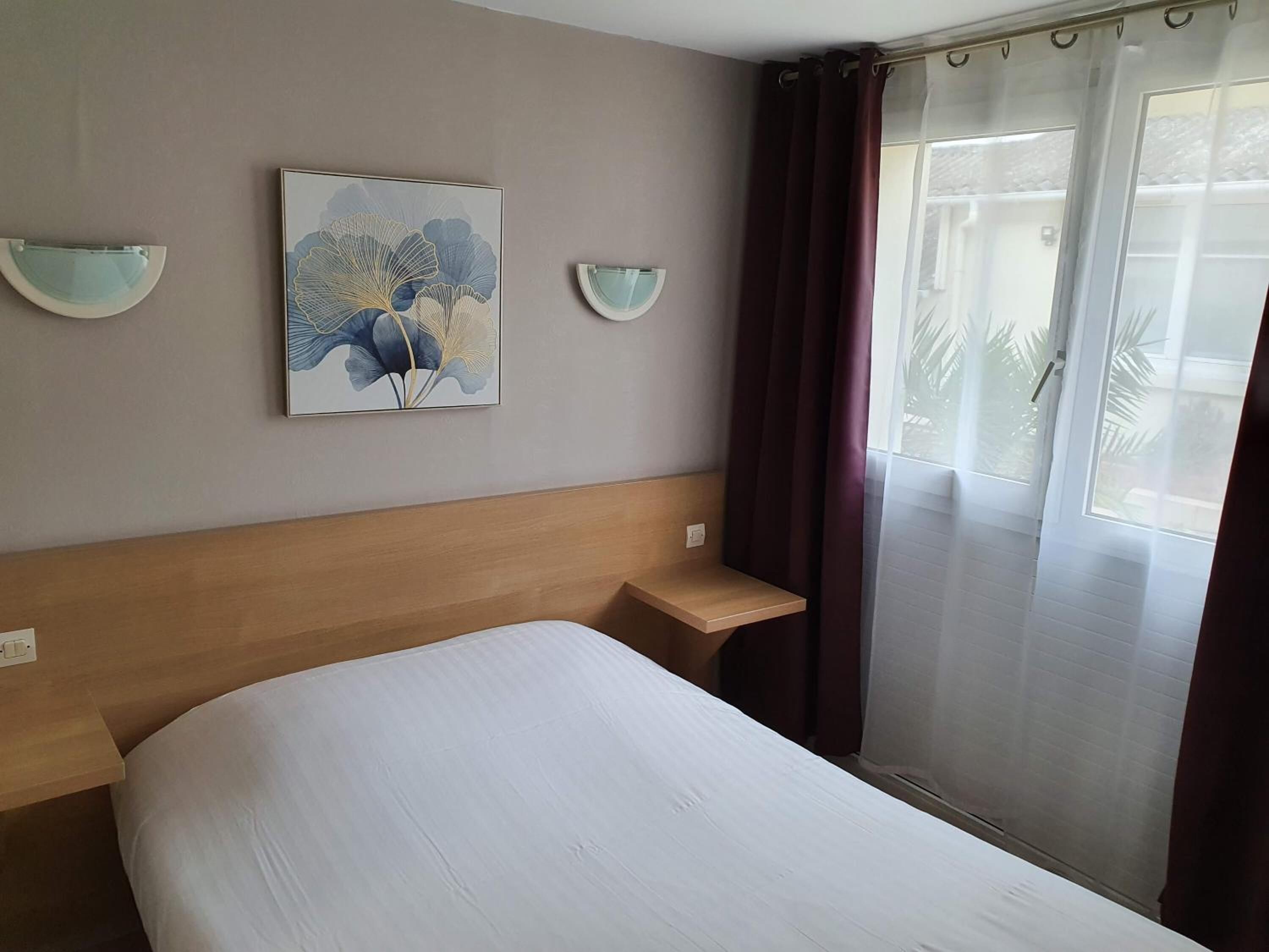 A hotel room with a bed, a headboard, a bedside table, a wall-mounted light fixture, and a window with curtains.
