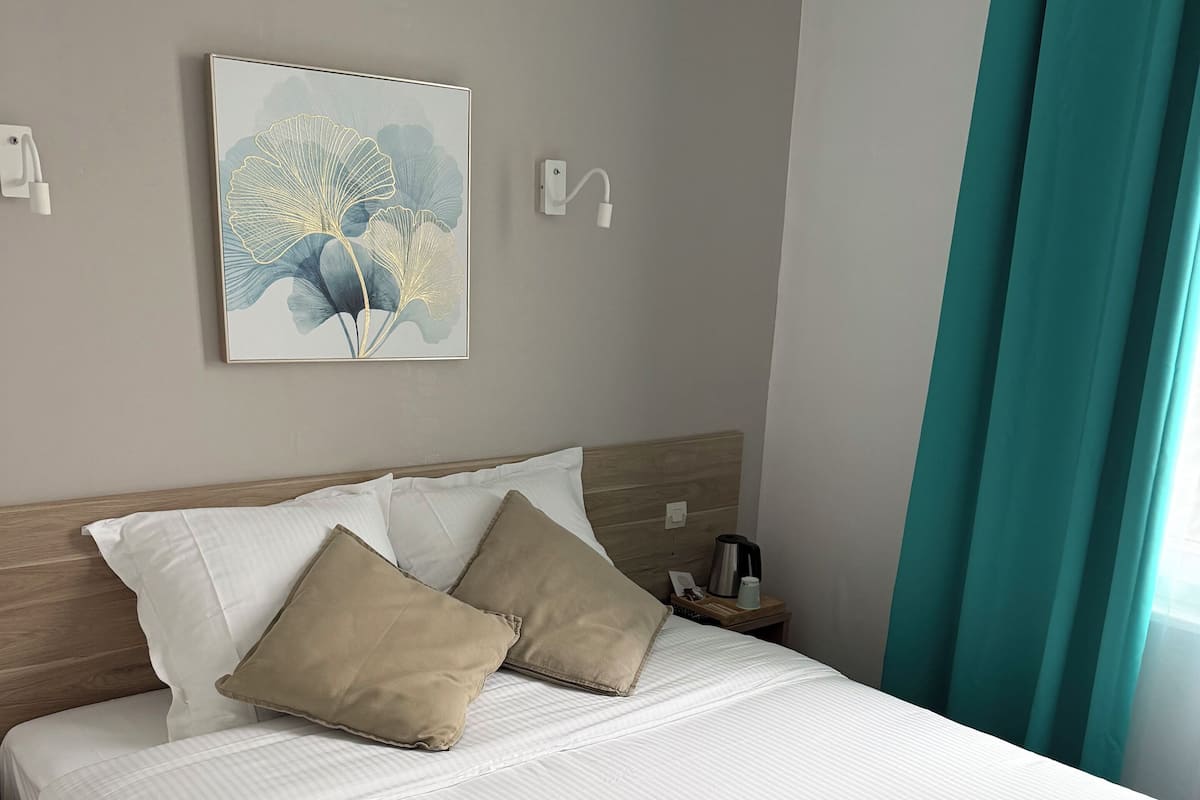 A neatly made bed with white linens and two beige pillows, a coffee maker on the nightstand, and a framed artwork on the wall.