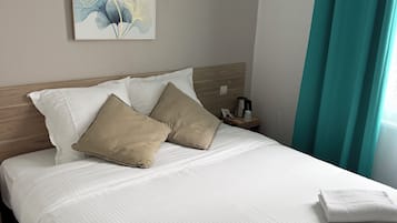 Una cama bien hecha con sábanas blancas y dos almohadas color beige, una cafetera sobre la mesita de noche y un cuadro en la pared.