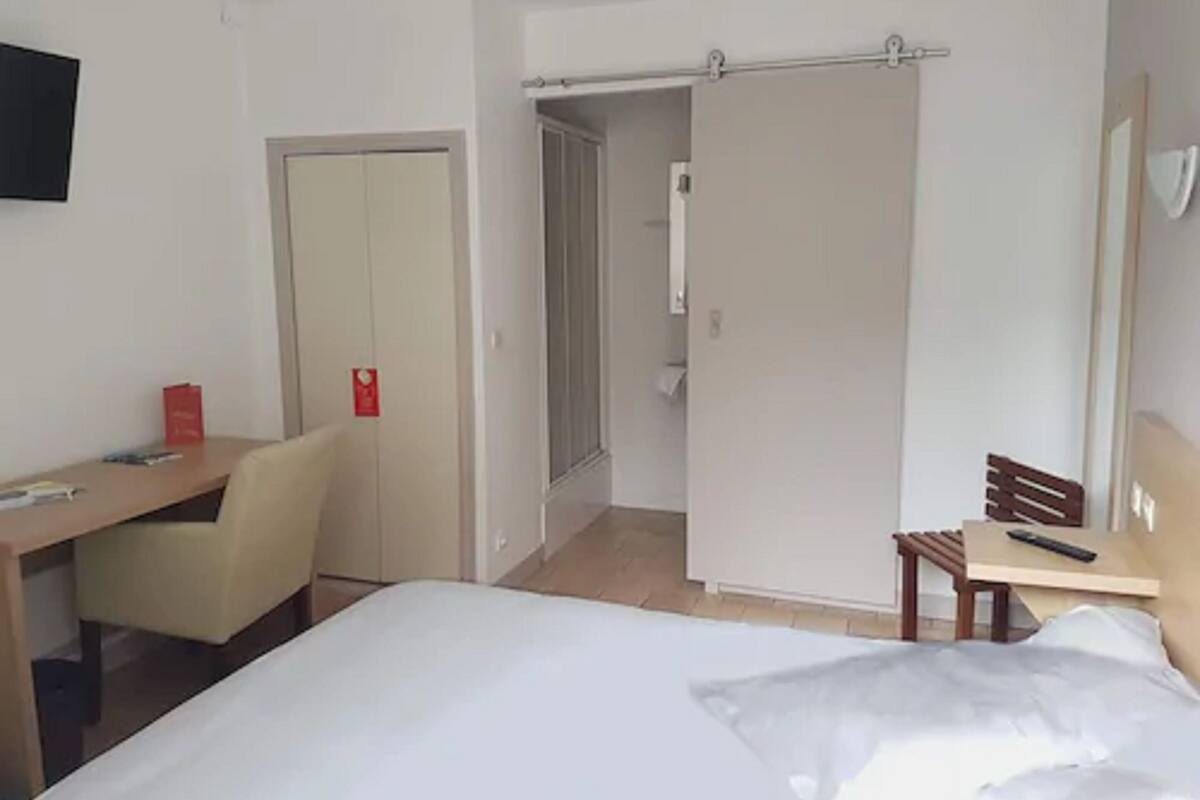A hotel room with a bed, a desk, a chair, a wardrobe, and a shower area.