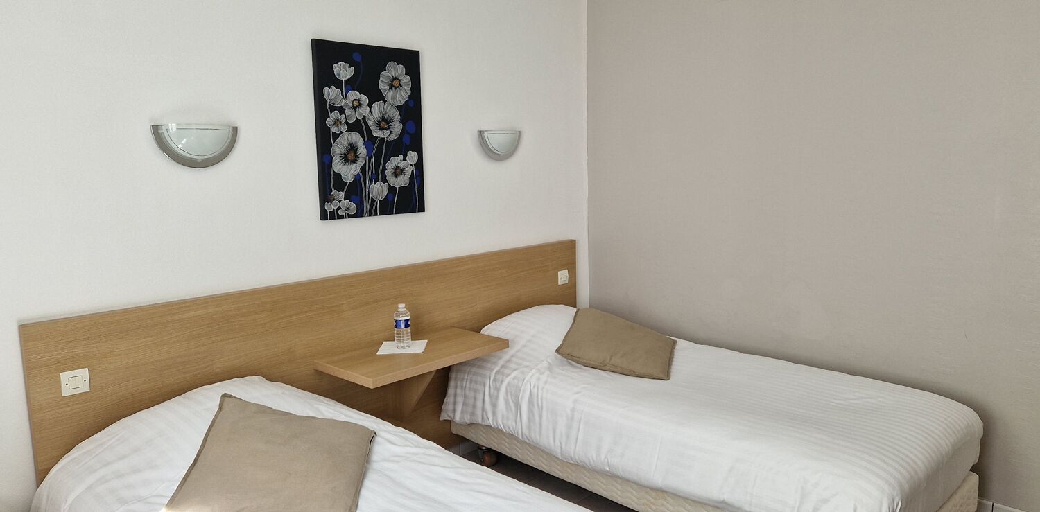 A hotel room with two single beds, a wooden headboard, a small bedside table with a water bottle, and a wall-mounted light fixture.