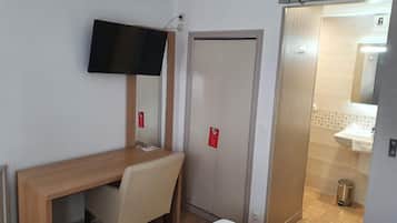 Una habitación de hotel con un escritorio de madera, una silla, un televisor en la pared, una puerta y un baño visible a través de una puerta abierta.