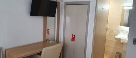 Hotelkamer met een houten bureau, een stoel, een aan de muur bevestigde televisie, een deur en een zichtbare badkamer door een open deur.