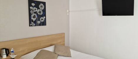 Habitación de hotel con cama, cabecera de madera, cuadro de flores, televisor de pantalla plana montado en la pared y mesita de noche con botella de agua, cafetera y control remoto.