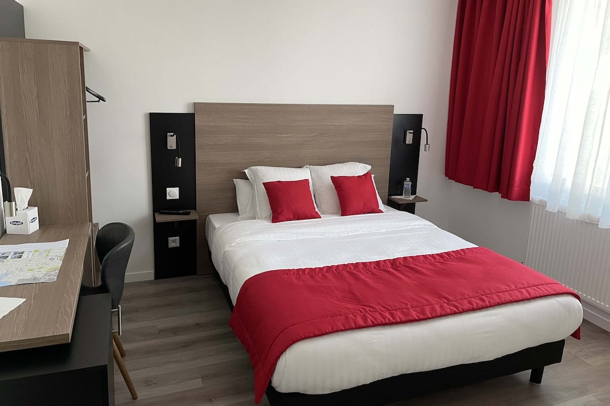 A hotel room with a bed, a desk, a chair, a wardrobe, and a window with red curtains.