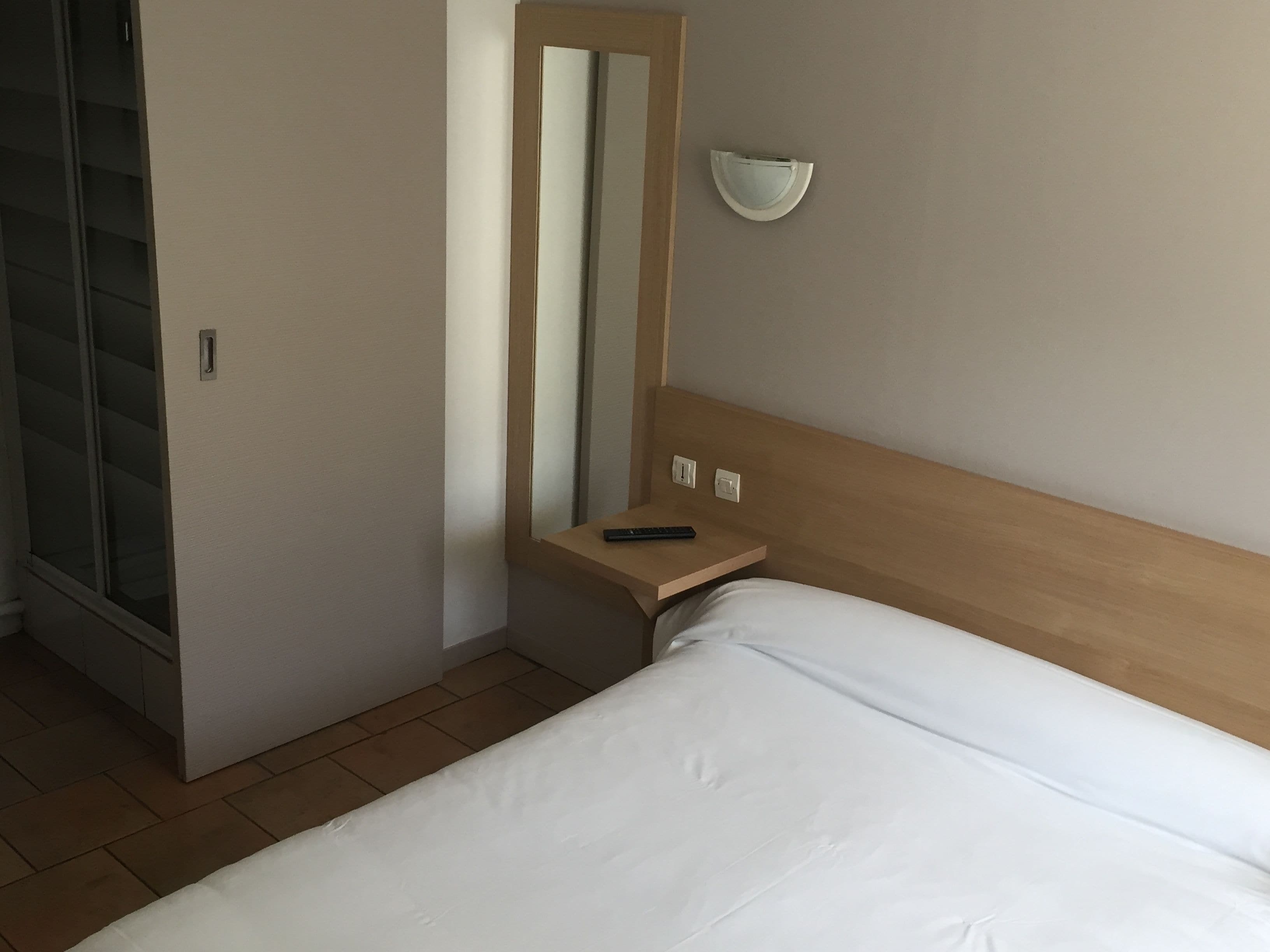 A hotel room with a bed, a bedside table with a remote control, a mirror, and a wall-mounted light fixture.
