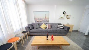 A modern living room with a grey sofa, wooden coffee table, and small stools. - YalaRent Designed 1BR Apartments - Families only (Eilat)