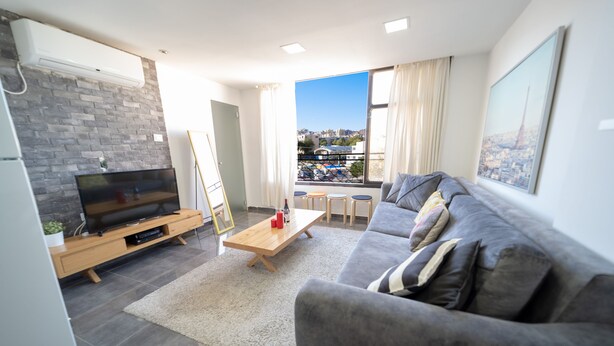 A modern living room with a grey sofa, a wooden coffee table, a flat-screen TV, and a city view through large windows. - YalaRent Designed 1BR Apartments - Families only (Eilat)