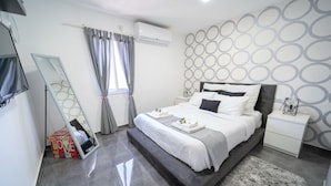A modern bedroom with a large bed, a bedside table, a mirror, and a grey patterned wall. - YalaRent Designed 1BR Apartments - Families only (Eilat)