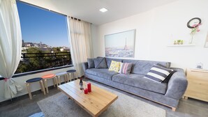 A modern living room with a grey sofa, a wooden coffee table, and a view of a cityscape through large windows. - YalaRent Designed 1BR Apartments - Families only (Eilat)