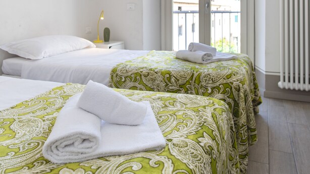 Quarto de hotel com duas camas, mesa de cabeceira com abajur e vista da cidade através de amplas janelas.