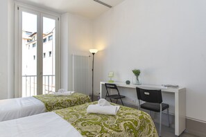 Habitación de hotel con dos camas, un escritorio, una silla, una lámpara y vistas a edificios a través del ventanal.