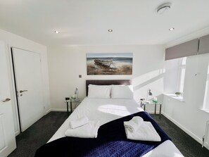 A neatly arranged bedroom with a bed, a painting of a boat on a beach, and a window providing natural light. - The Barbican Penthouse (Plymouth)
