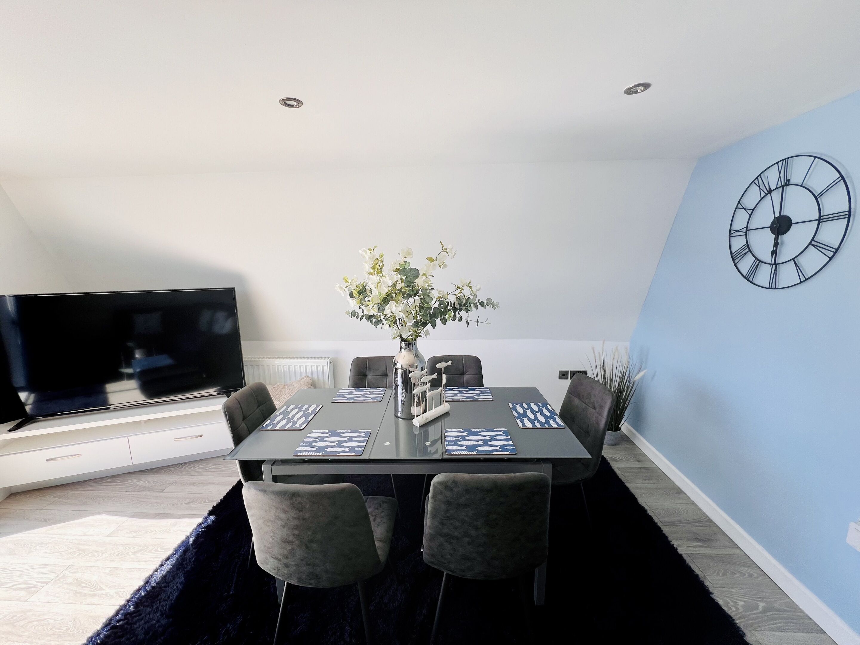 A modern dining area with a black table, grey chairs, a vase with white flowers, and a wall clock.