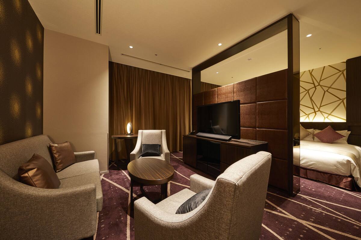 Une chambre d’hôtel avec un canapé, un fauteuil et une petite table ronde. On y trouve également une télévision et un lit avec une tête de lit.