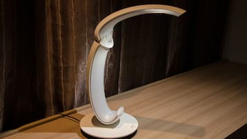 En modern, böjd skrivbordslampa med en vit bas och en metallarm, placerad på en träyta mot en mörk draperad bakgrund.